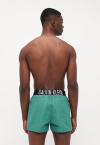 Zelene kupaćice s crnim pojasom na kojem je u bijelom ispisano "CALVIN KLEIN". Materijal izgleda glatko; kupaćice imaju stražnji džep.