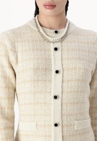Cardigan en crème texturé avec des boutons noirs, deux poches et un collier en perles et métal superposé au niveau du cou.