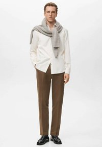 Camisa de rayas blancas, bufanda gris de punto sobre los hombros, pantalones marrones, zapatos negros. Diseño sencillo con un corte entallado y colores apagados.