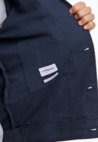 Binnenkant van een marineblauw jack met een platte zak en merklabel. De stof is soepel, met gestikte naden en twee kleine uitsparingen aan de onderkant.