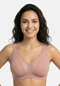 Uśmiechnięta kobieta o ciemnych kręconych włosach, ubrana w jasnoróżowy biustonosz bez fiszbin, stojąca przodem na tle jednolitego jasnego tła.