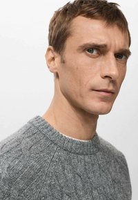 Szary sweter z dzianiny warkoczowej z okrągłym dekoltem, ozdobiony teksturowanymi wzorami i prążkowanymi detalami, noszony przez mężczyznę z krótkimi włosami na neutralnym tle.