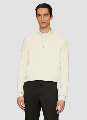 Cream zip-neck pullover sweater with long sleeves, soft texture, and a fitted silhouette, paired with dark trousers.