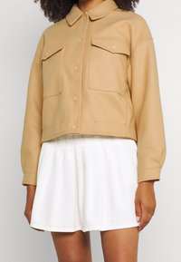 Beżowa krótka kurtka z zapięciem na guziki, dwoma kieszeniami na piersi i z kołnierzykiem. Noszona na białej plisowanej spódnicy. Gładka faktura.