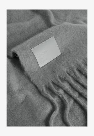 Šedý pletený šátek vyrobený z jemného, texturovaného materiálu s třásněmi na okrajích a koženou etiketou s embosovaným nápisem "přátelé jako tiše".