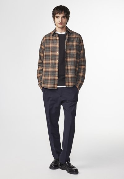 Men's brown and grey plaid flannel shirt, buttoned, over a dark sweater, paired with navy trousers and black loafers. Simple design.