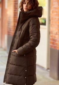 Manteau rembourré marron avec un col montant, fermeture éclair et poches latérales. Tissu texturé avec des coutures matelassées et des détails en pression métallique.