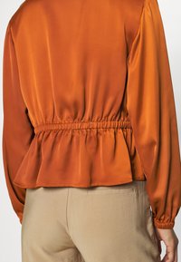 Rust-orange satin bluse med peplum nederst og elastisk talje. Lange ærmer med samlede manchetter. Neutrale beige bukser nederst.