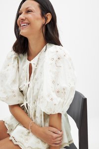 Robe de couleur claire avec des manches bouffantes, broderie florale et un col à cravate. Présente des motifs délicats et un ourlet volé.