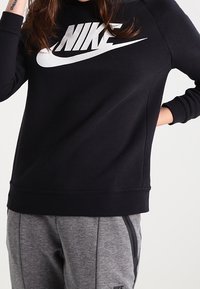 Czarna bluza Nike z dużym białym logo. Posiada okrągły dekolt i ściągacze na rękawach. Noszona z szarymi sportowymi spodniami.