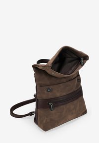 Mochila de cuero marrón con una tapa plegable, bolsillo frontal con cremallera y correa ajustable. Textura suave con sutiles acentos metálicos.