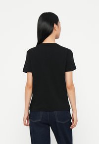 Melna kokvilnas T-krekls ar apaļu kakla izgriezumu un īsām piedurknēm, kombinēts ar tumši zilām džinsa biksēm. Aizmugure ir gluda bez redzamiem logotipiem.