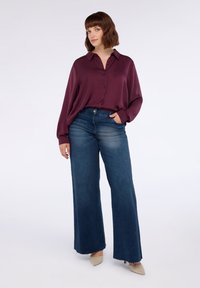 Camicia button-up bordeaux, vestibilità ampia, con maniche lunghe. Jeans blue a gamba larga, lavaggio medio, e tacchi beige a punta. Sfondo bianco semplice.