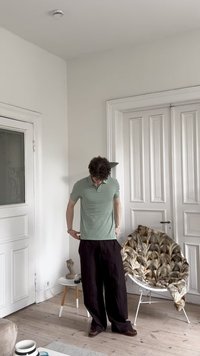 Camisa polo verde claro com colarinho, combinada com calças largas pretas. O ambiente apresenta portas brancas e uma cadeira de padrão suave.