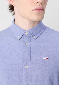 Camisa de botón de color púrpura claro con cuello, que presenta pequeños botones y una insignia con logotipo en el lado izquierdo del pecho. Tejido suave y texturizado.