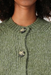 Dettaglio ravvicinato di una persona che indossa un cardigan chunky verde con due bottoni marmorizzati allacciati vicino al collo e al petto.