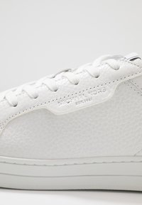Zapatilla blanca hecha de cuero texturizado, con punta redonda, cordones blancos y un sutil detalle de marca en el lateral. Suela de goma.