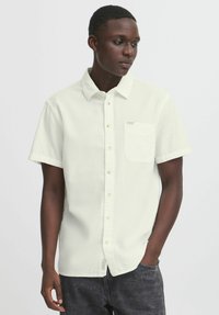 Camisa de manga corta, de color crema claro, con un patrón texturizado, cierre de botones en la parte delantera y un bolsillo en el pecho. Marca sutil en el área del bolsillo.