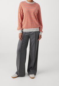 Maglione a righe arancione e grigio con maniche lunghe e polsini a coste, abbinato a pantaloni grigi a gamba larga. Scarpe bianche completano l'outfit.