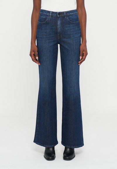 Personne portant un jean évasé bleu foncé, un haut blanc et des bottes noires à bout pointu, se tenant devant un fond clair.