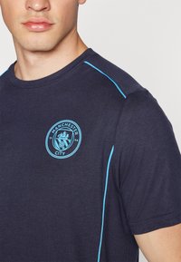 Jūras spēku zila kokvilnas T krekls ar gaiši zilu apdari plecu daļās un Manchester City logo uz krūtīm. Mīksts, ikdienas piegriezums.