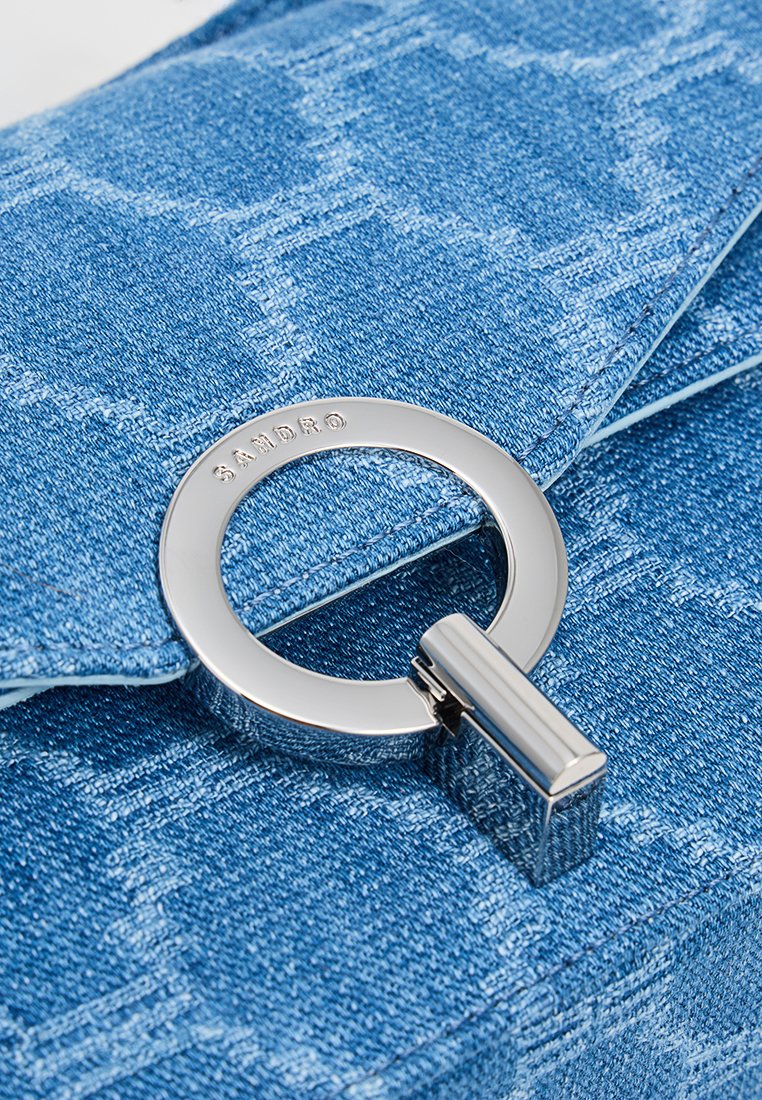 Blue denim bag with a textured finish, featuring a round silver metal clasp with "SANDRO" engraved, and contrasting light blue stitching.