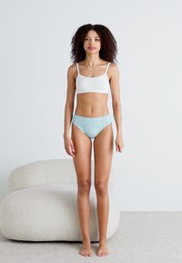 Witte katoenen bralette en lichtblauwe kanten slipjes tonen een glad ontwerp. Model staat blootsvoets tegen een neutrale achtergrond.