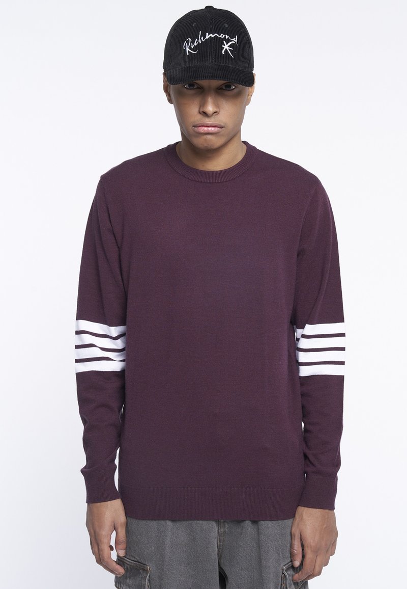 Maglione lungo color bordeaux con strisce bianche sulle maniche. Indossato con pantaloni corti grigi e un berretto nero con testo del logo bianco.