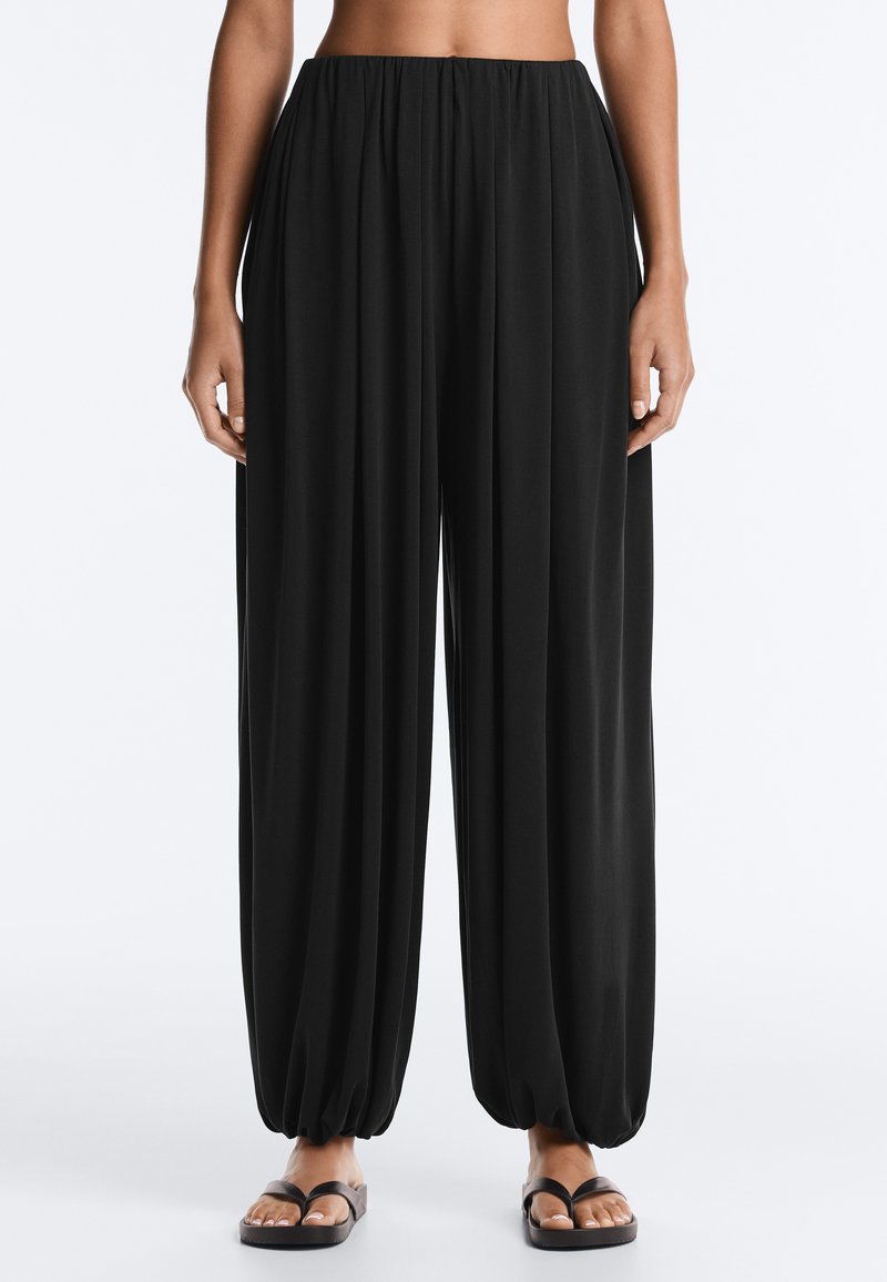 Femme portant un pantalon noir ample avec des poignets froncés et des sandales noires, debout devant un fond clair uni.