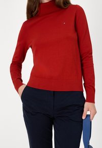 Punainen poolopaita, joka on valmistettu pehmeästä materiaalista ja siinä on pitkät hihat sekä vartalonmyötäinen malli, jossa on pieni logotunnistus rinnassa. Laivastonsiniset housut.