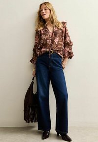 Bruine blouse met bloemenpatroon en ruche mouwen, gecombineerd met hoge donkere denim wijde spijkerbroek en een zwart handtas met franjes.