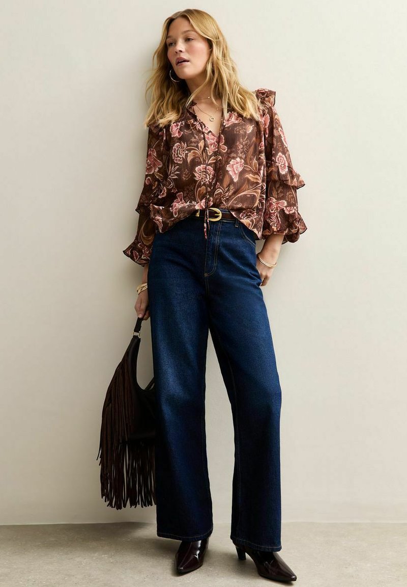 Bruine blouse met bloemenpatroon en ruche mouwen, gecombineerd met hoge donkere denim wijde spijkerbroek en een zwart handtas met franjes.
