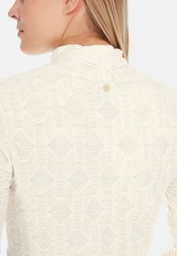 White top with a textured geometric pattern, featuring a small gold accent button at the back and a ruffled neckline.