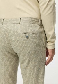 Világos színű, texturált nadrág, finom kockás mintával, hátul egy kék gombbal rögzített zsebből és szabott derékból áll.