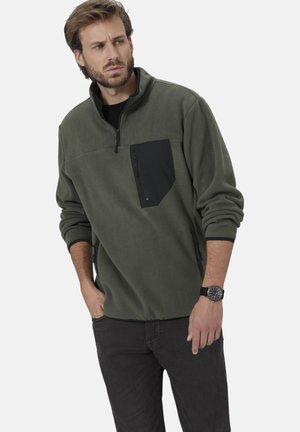 Grüner Fleece-Pullover mit einer schwarzen Reißverschluss-Brusttasche, hohem Kragen, langen Ärmeln und strukturiertem Stoff. Kombiniert mit dunklen Jeans und einer Uhr.