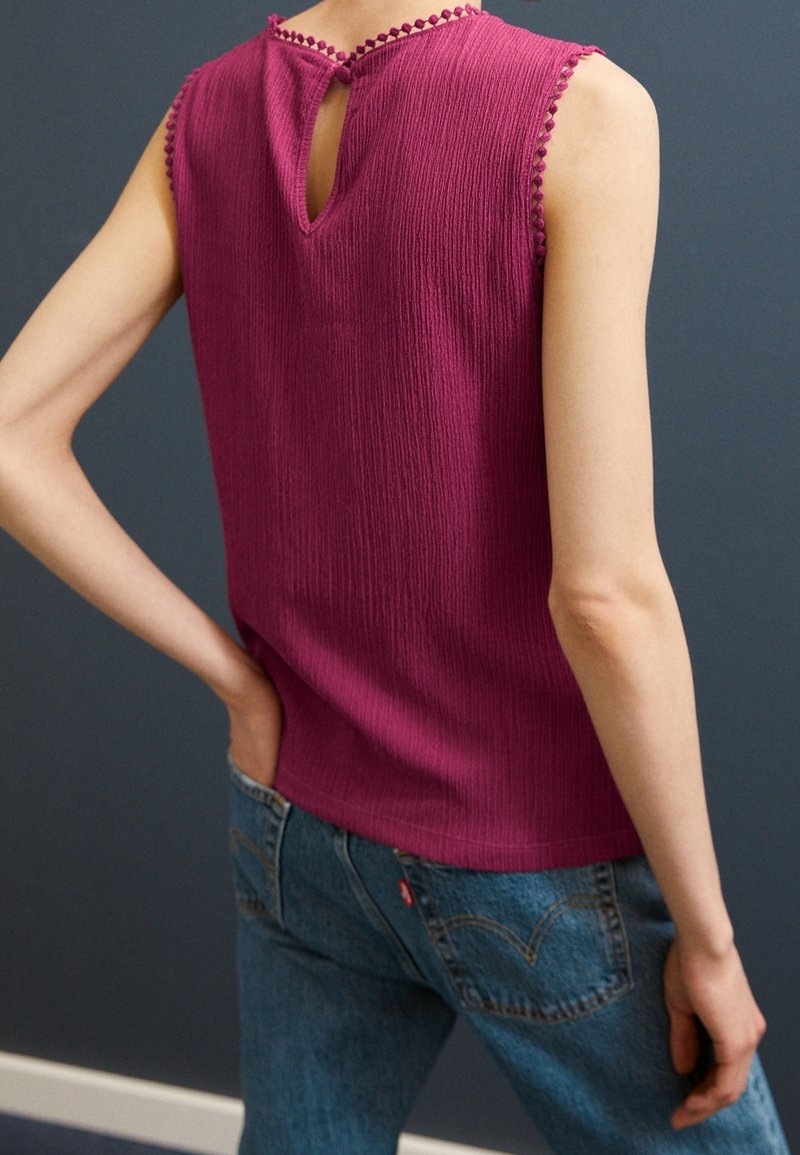 Fuchsia sleeveless top with textured fabric, keyhole back detail, and pom-pom trim. Paired with blue denim jeans.