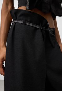 Zwarte broek met wijde pijpen en een gestructureerde tailleband, voorzien van een contrasterend plaid strikdetail. Gladde stof met een ontspannen pasvorm bij de taille.