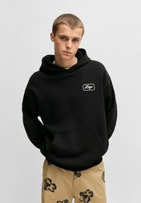 Felpa nera con tasca frontale e logo ricamato. Abbinata a pantaloni corti beige con motivo floreale. Tessuto morbido.