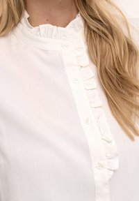 Blouse blanche à col volanté et devant boutonné, portée par une personne aux longs cheveux blonds sur l'épaule.