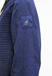 Chaqueta deportiva azul marino que presenta una textura acolchada en un lado, con un tejido suave en el otro. Incluye un parche con el logo de Adidas en la manga.