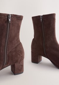 Hnedé semišové členkové topánky so štvorcovým podpätkom, vybavené bočným zipsom a prešívanými detailmi pre textúru a štruktúru.