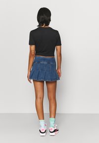 Ung person som står vänd bort ifrån kameran, klädd i en svart crop top, blå plisserad denimkjol, tie-dye strumpor och vita sneakers med rosa detaljer.