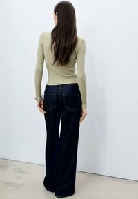 Top a maniche lunghe a coste oliva abbinato a jeans svasati blu scuro. I jeans presentano tasche posteriori e dettagli di cucitura bianca a contrasto.