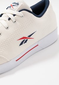 Zapatilla de lona Reebok con un upper en color crema, acentos en azul marino, logo rojo y suela de goma blanca. Presenta cordones planos y detalles cosidos.