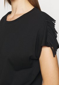 Svart bomulls t-shirt med korta ärmar som har texturerad fransdetalj längs axlarna. Enkel rund halsringning, mjukt tyg.