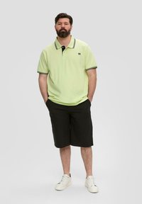 Jasnozielona koszulka polo z czarnym kołnierzykiem i mankietami, połączona z czarnymi szortami i białymi trampkami. Koszulka ma małe logo na piersi.