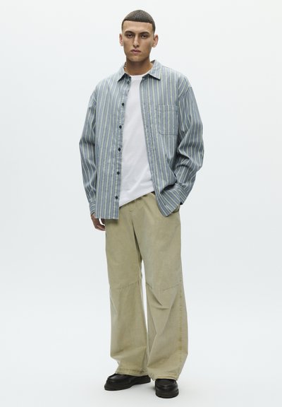 Egy férfi áll egy egyszerű háttér előtt, kék csíkos gombos inget, fehér pólót, bézs színű, széles szárú nadrágot és fekete cipőt viselve, kezét a zsebében tartva.