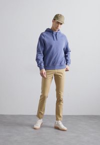 Sweatshirt azul claro com bolso frontal, calças beigas, e ténis brancos com solas de borracha. Modelo a usar um chapéu bege, de pé contra um fundo neutro.