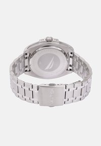 Kovinska wristwatch iz nerjavnega jekla z povezavnim pasom, ki ima okroglo ohišje in prozorno zadnjo stran, na kateri je prikazana znamka Nautica. Srebrni odtenek skozi celotno zasnovo.