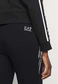 Černé sportovní oblečení s bílým logem EA7. Má těsný střih, měkkou texturu a kontrastní pruhy podél rukávů a po stranách kalhot.